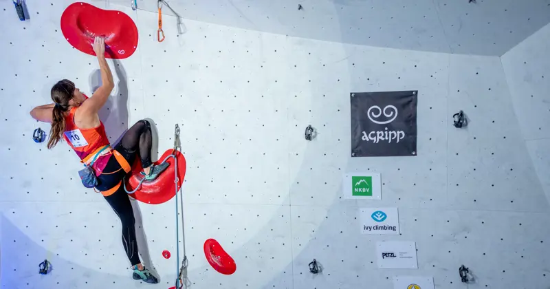 Een vrouw met een klimgordel beklimt een indoorklimwand, terwijl ze zich vasthoudt aan grote rode grepen. Ze neemt deel aan een wedstrijd, zoals blijkt uit haar startnummer en de sponsorbanieren op de witte klimwand achter haar.